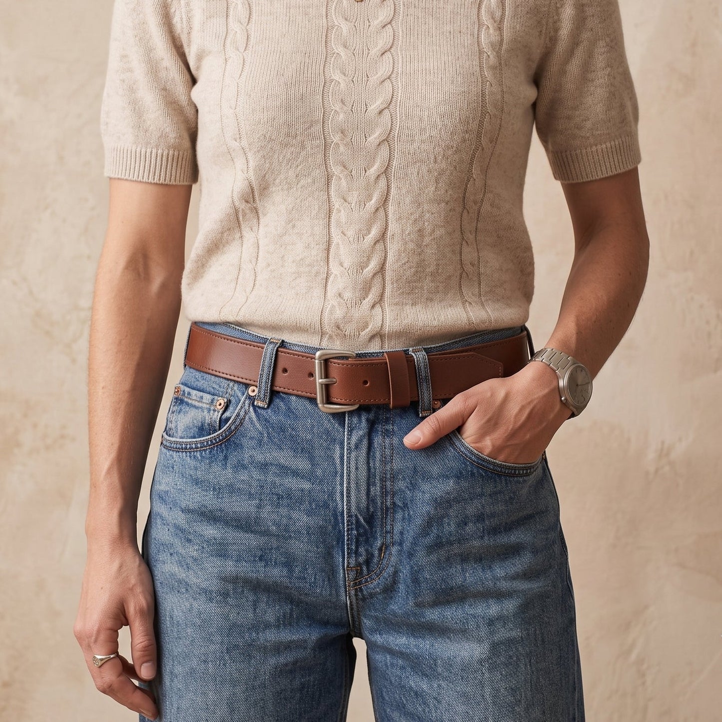 A woman wearing a beige short-sleeve cable-knit sweater, blue jeans, and a brown leather belt, standing against a textured beige wall.