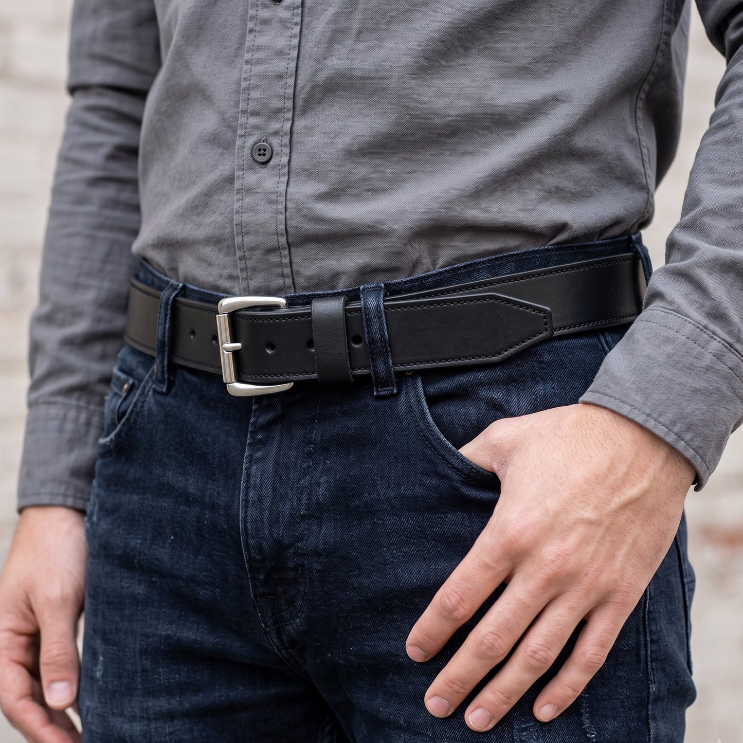 A close-up of a man wearing a black leather belt with a silver-tone buckle, tucked into dark blue jeans, paired with a gray button-down shirt.