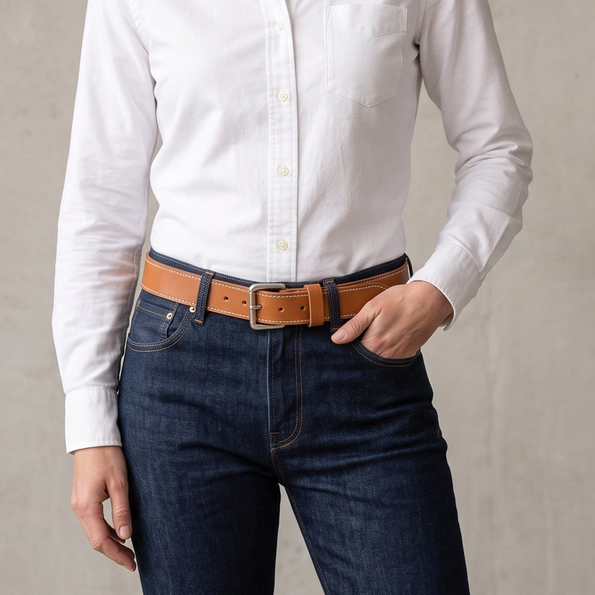 A woman wearing a white long-sleeve button-down shirt tucked into dark blue jeans with a brown leather belt, standing against a textured light gray wall.