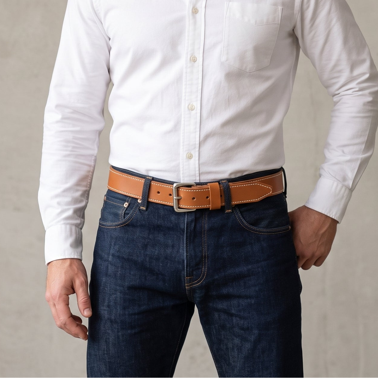 A man wearing a white long-sleeve button-down shirt, dark blue jeans, and a brown leather belt, standing against a textured gray wall.