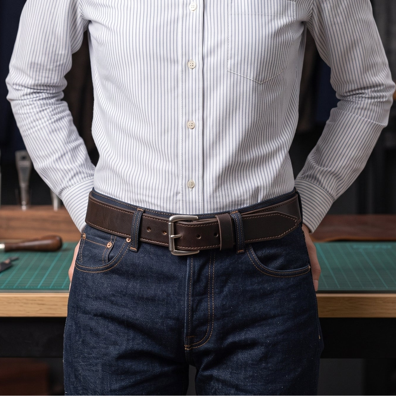 A woman wearing a light blue and white vertically striped button-down shirt, paired with a brown belt and dark blue jeans, standing in a workshop environment.